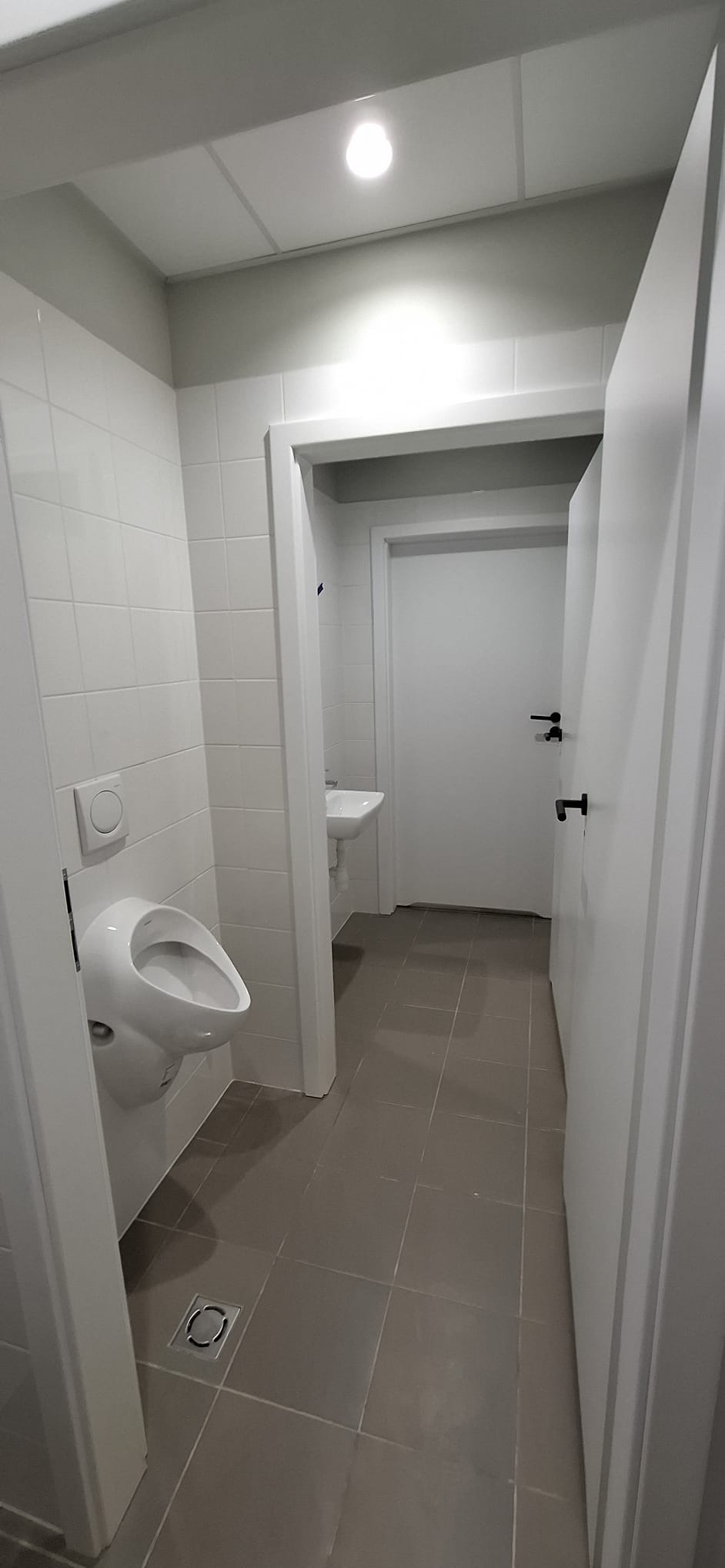 Nowoczesna toaleta publiczna z białymi płytkami, wiszącym pisuarze i szarą podłogą z płytek. Widoczne drzwi i umywalka w tle. Minimalistyczny design.