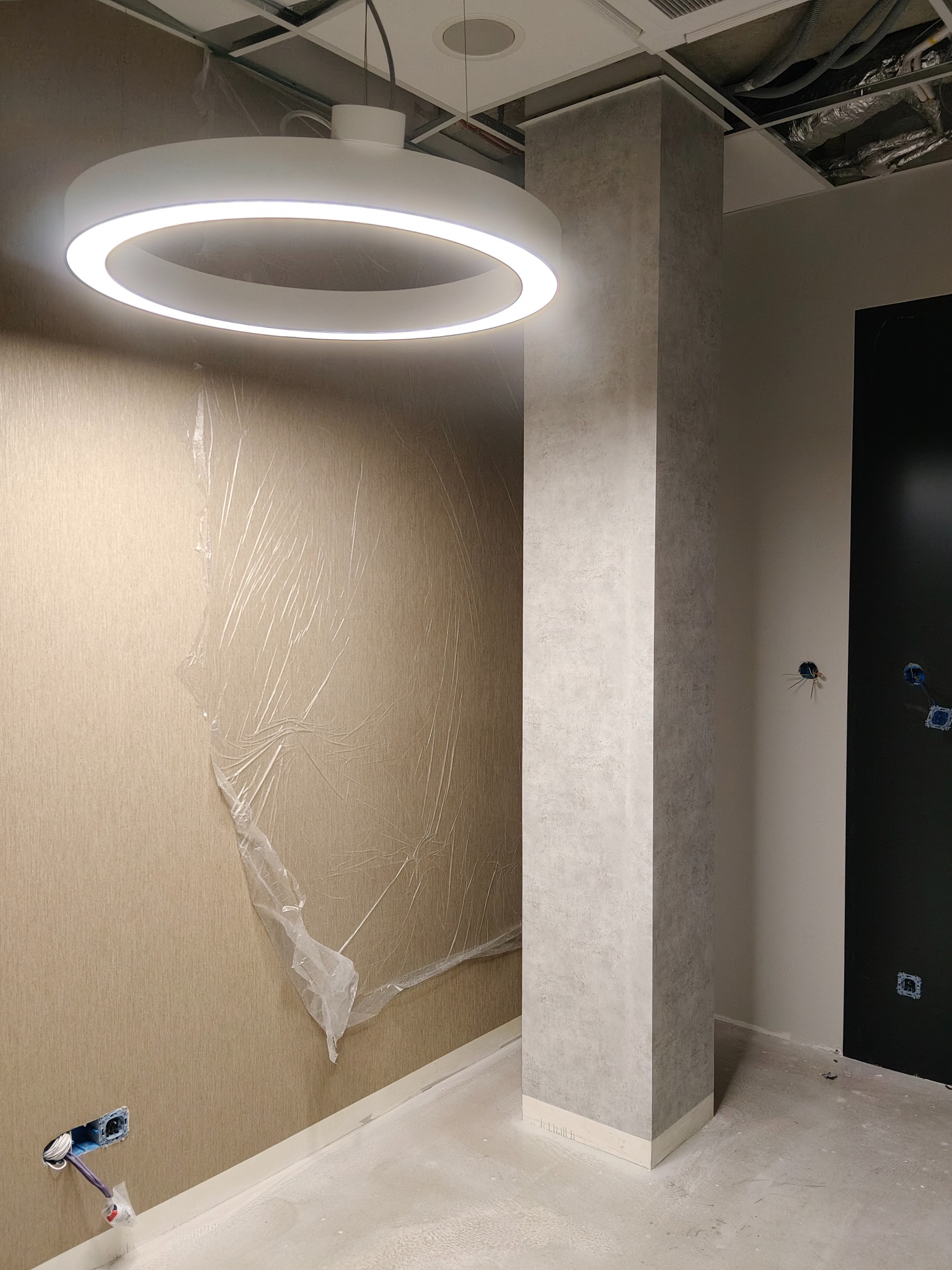 Wnętrze w trakcie wykańczania: kolumna obłożona płytami, ściana zabezpieczona folią, okrągła lampa sufitowa. Widoczne gniazdka elektryczne i surowy sufit.