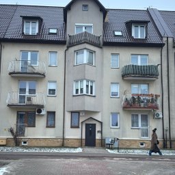 KUPREM Sp. z o.o. - Wielorodzinny budynek z ciemnym, skośnym dachem z lukarnami i balkonami z ozdobnymi balustradami. Osoba przechodzi przez przejście dla pieszych.