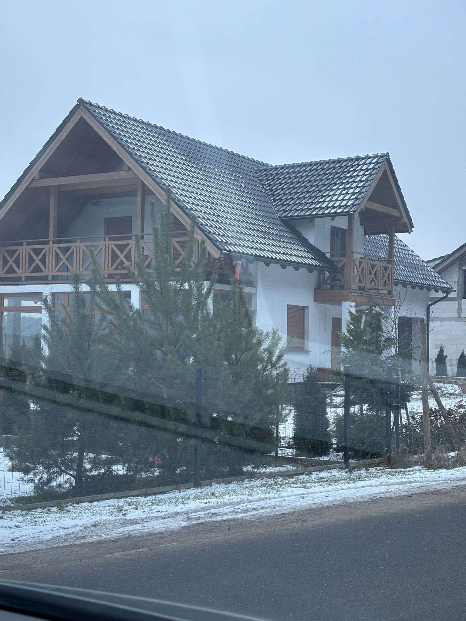 Dwukondygnacyjny dom z ciemną dachówką i drewnianymi balkonami, widziany zza ogrodzenia. Śnieg na ziemi, pochmurne niebo.
