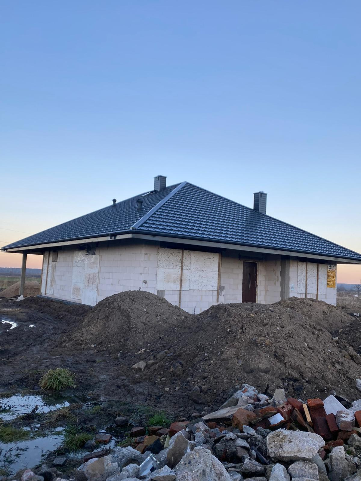 Dom w trakcie budowy z ciemnym, metalowym dachem. Widoczne bloczki i stosy ziemi. Dach pokryty blachodachówką, z kominami. Budynek w stanie surowym.