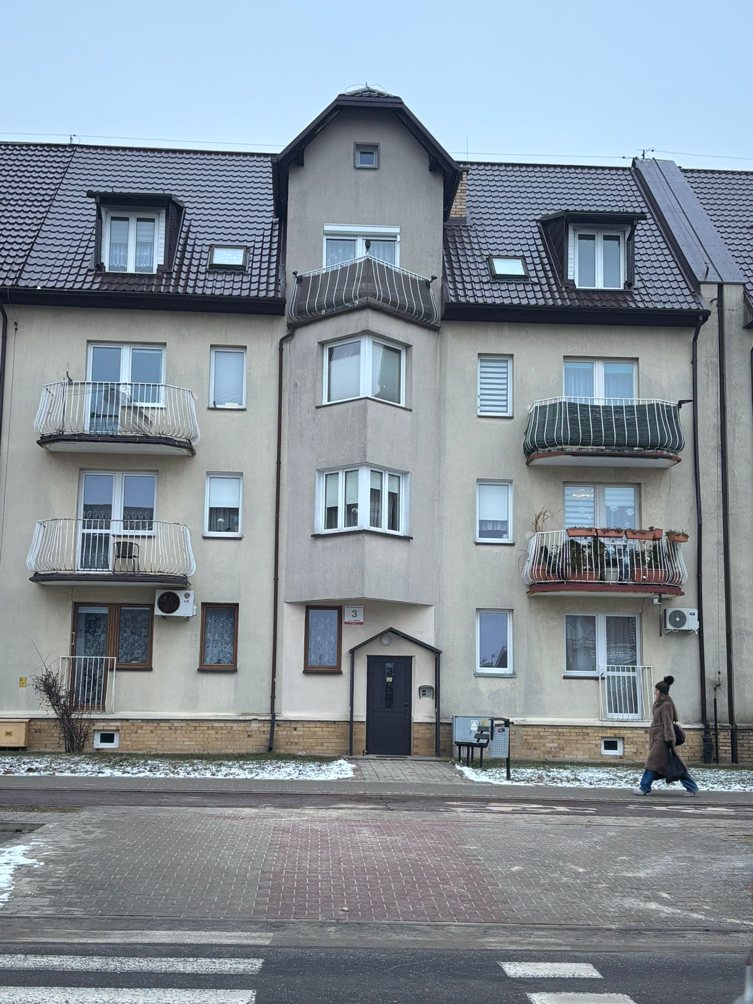 Wielorodzinny budynek z ciemnym, skośnym dachem z lukarnami i balkonami z ozdobnymi balustradami. Osoba przechodzi przez przejście dla pieszych.