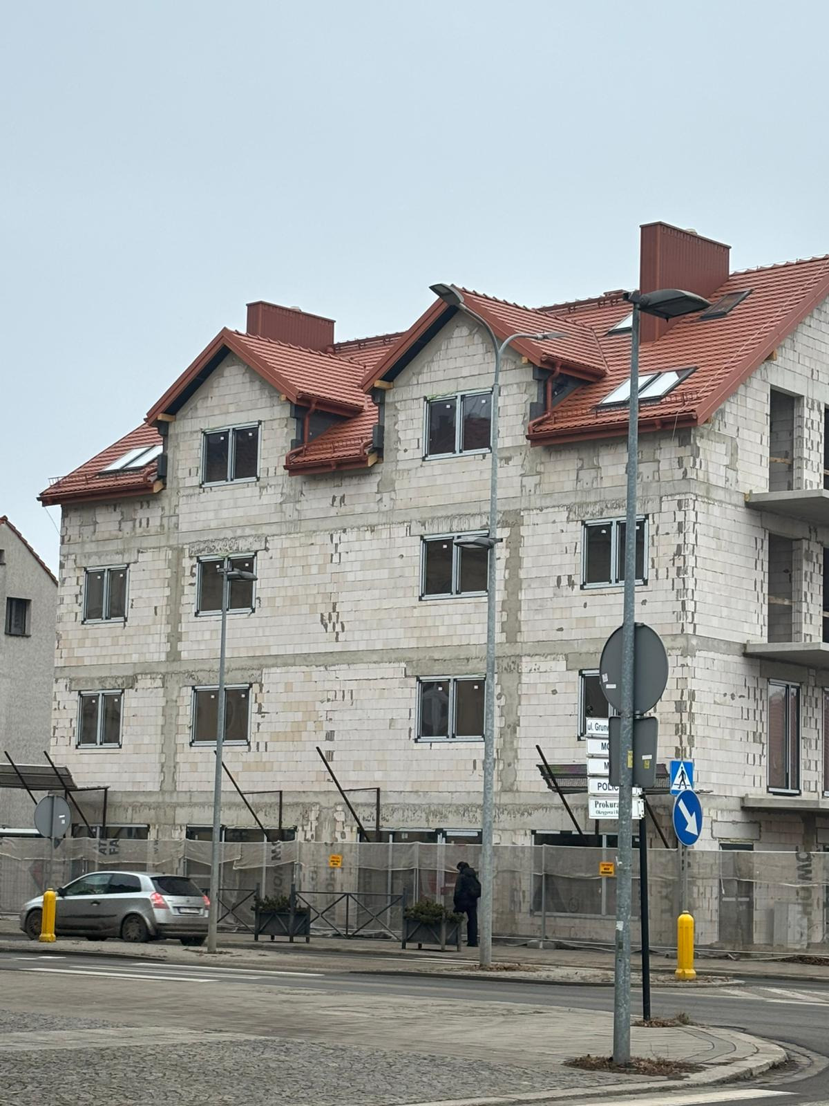 Budynek w trakcie budowy z czerwonym dachem i oknami. Widoczne rusztowania i ulica z chodnikiem. Pochmurne niebo w tle.