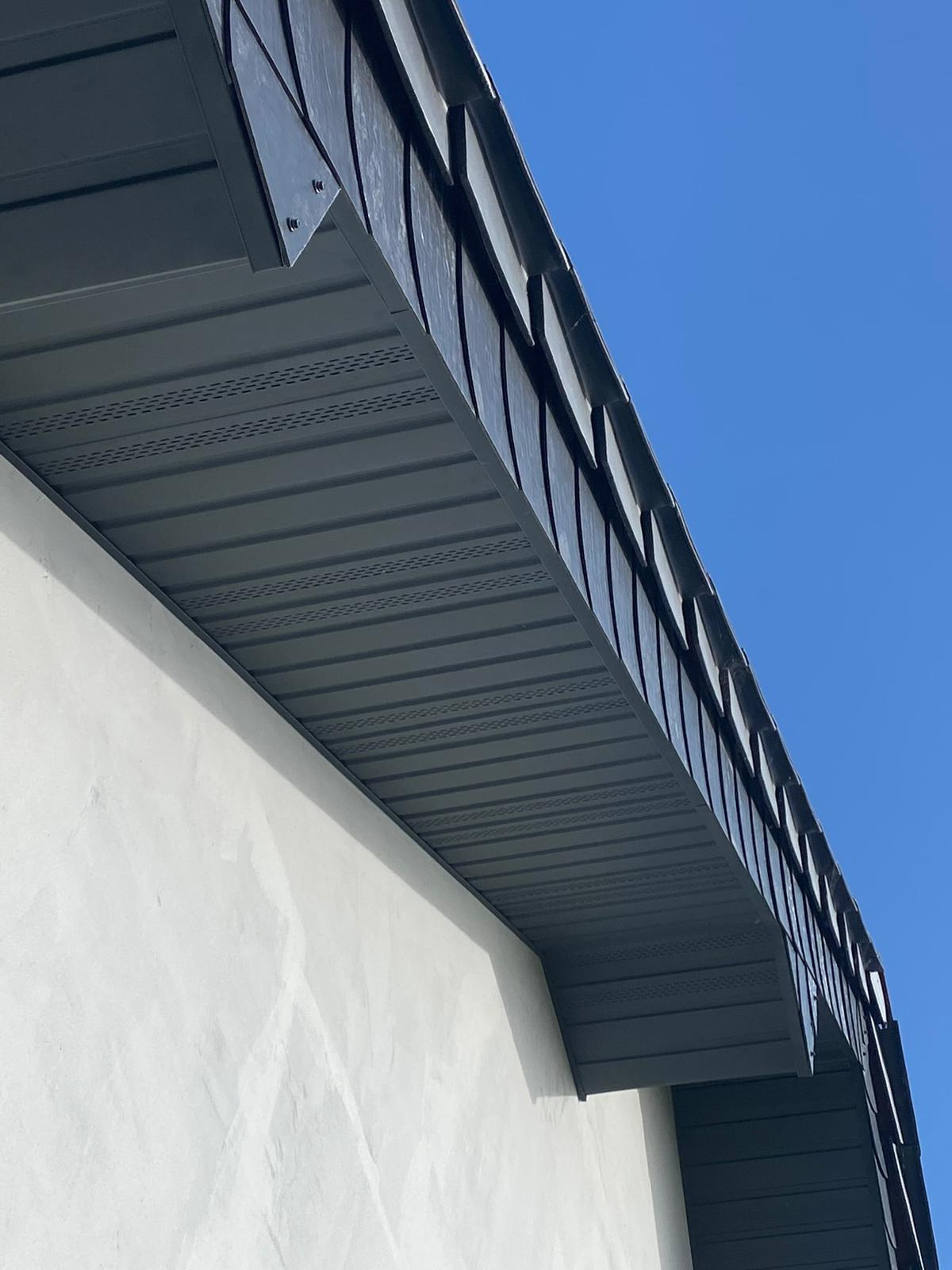 Szare podbitki dachowe z widocznymi otworami wentylacyjnymi, zamontowane przy białej ścianie budynku na tle błękitnego nieba. Ujęcie z dołu.