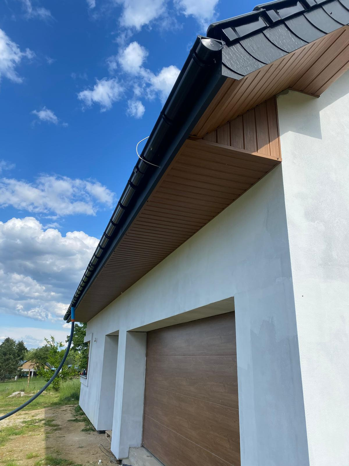 Nowoczesny dom z ciemną dachówką i orynnowaniem, jasna elewacja i drewniana podbitka. Widok z dołu na fragment budynku w słoneczny dzień.