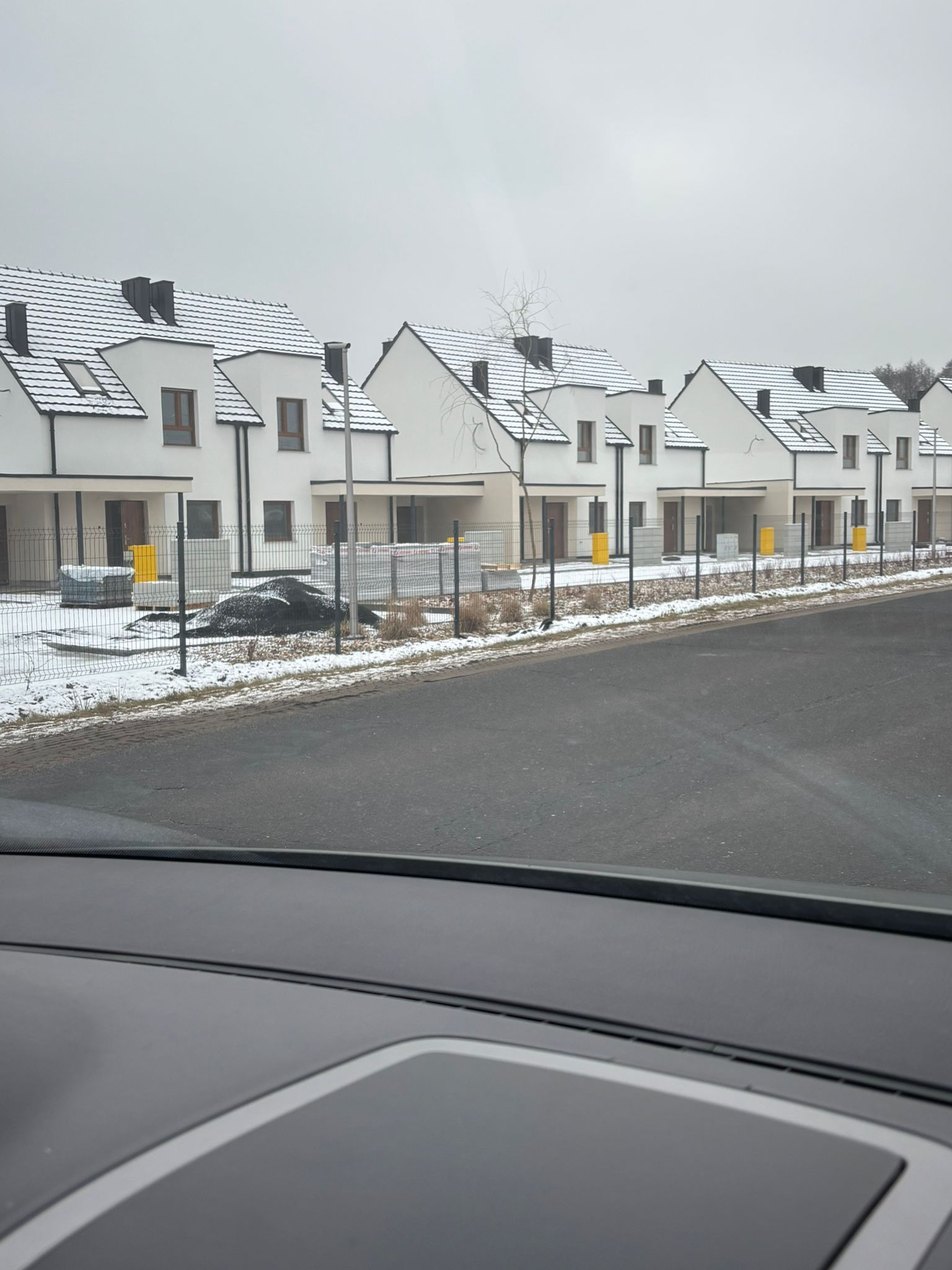 Rząd nowych, białych domów z ciemnymi dachami pokrytymi śniegiem, widzianych z perspektywy ulicy w pochmurny dzień. Częściowo widoczny kokpit samochodu.