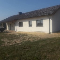 Firma remontowa Seb Bud - Biały dom jednorodzinny z ciemną dachówką i szarymi oknami w słoneczny dzień. Widoczny fragment trawnika i przyczepa z łodzią pod plandeką.