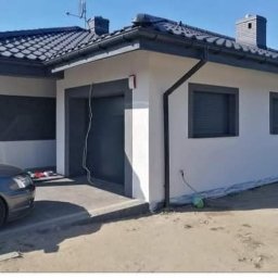 Firma remontowa Seb Bud - Biały dom z szarymi akcentami: dach, okna, brama garażowa. Widoczny fragment samochodu i teren wokół domu. Dom po wykończeniu, w słoneczny dzień.