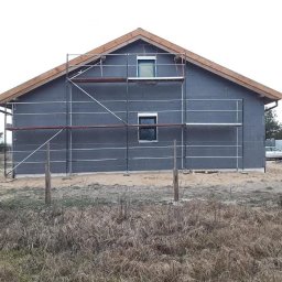 Firma remontowa Seb Bud - Ocieplony budynek z szarego bloczka, z drewnianą konstrukcją dachu i rusztowaniem. Widoczne okna i otoczenie budynku: trawa, drzewa, samochód.