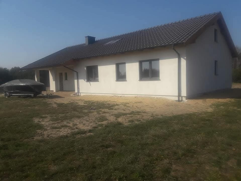 Biały dom jednorodzinny z ciemną dachówką i szarymi oknami w słoneczny dzień. Widoczny fragment trawnika i przyczepa z łodzią pod plandeką.