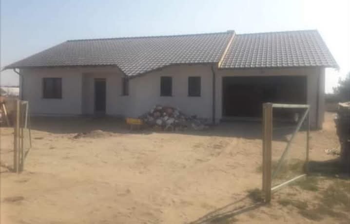 Parterowy dom jednorodzinny z garażem w stanie surowym zamkniętym, z szarą dachówką, na piaszczystej działce. Widoczne materiały budowlane i ogrodzenie.