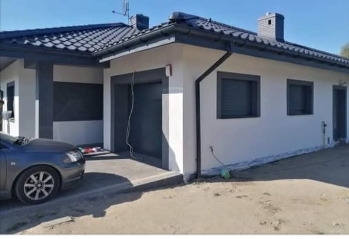 Biały dom z szarymi akcentami: dach, okna, brama garażowa. Widoczny fragment samochodu i teren wokół domu. Dom po wykończeniu, w słoneczny dzień.