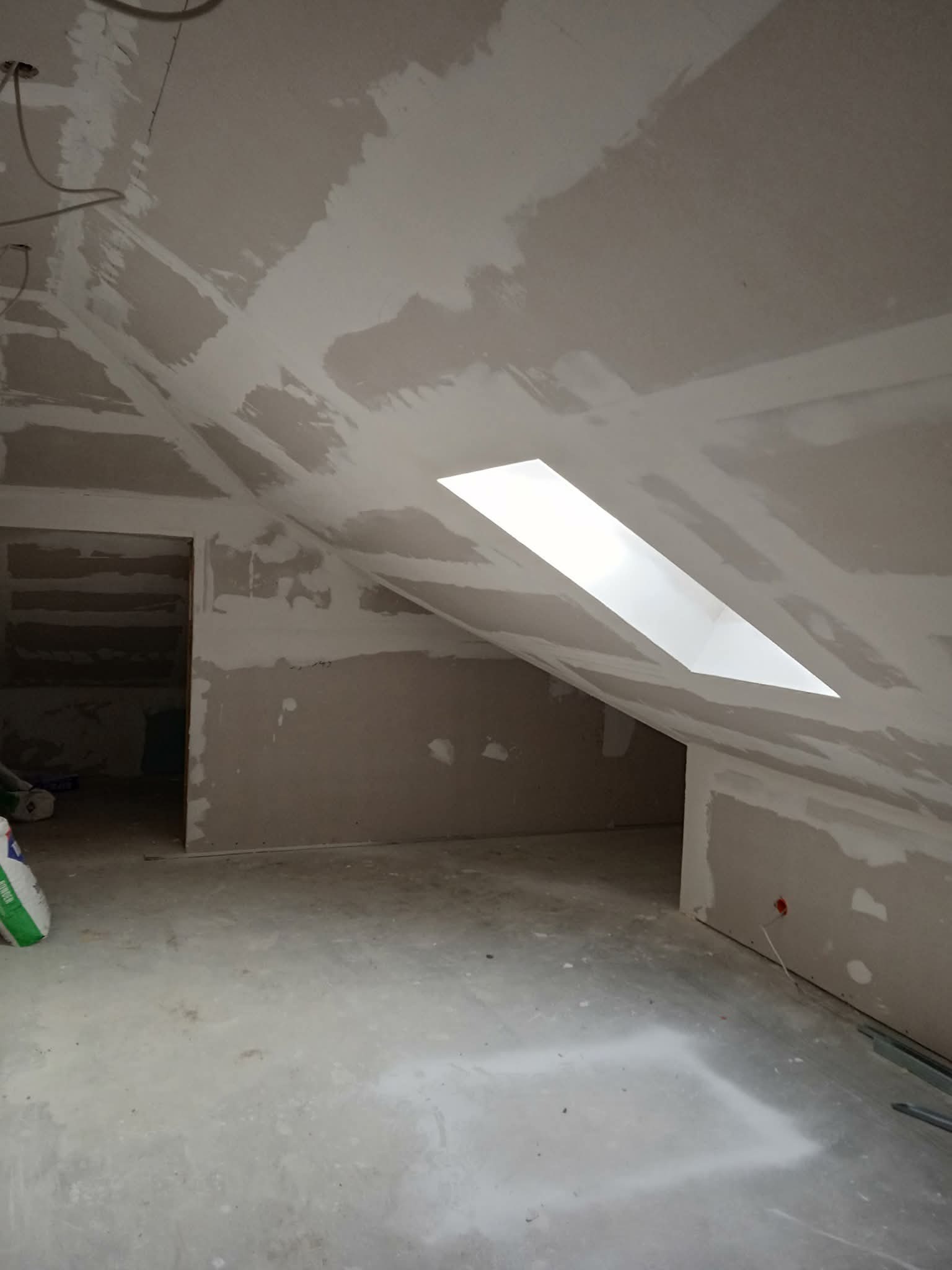 Poddasze po szpachlowaniu i montażu płyt gipsowo-kartonowych, widoczne okno dachowe i przygotowane ściany do dalszych prac wykończeniowych. W tle widoczne wejście do pomieszczenia.