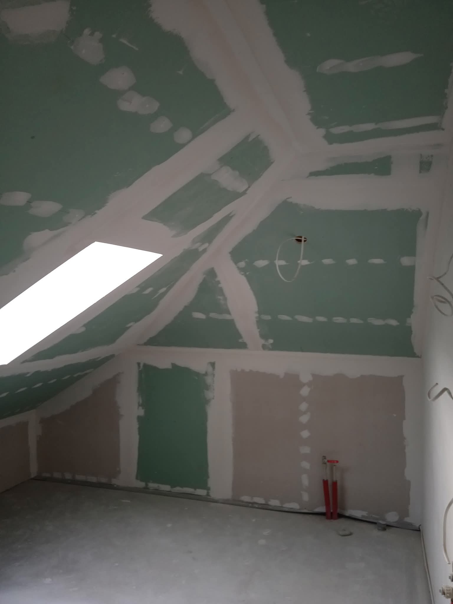 Poddasze z oknem dachowym w trakcie prac wykończeniowych: płyty gipsowo-kartonowe na skosach, szpachlowane łączenia, widoczne instalacje. Surowy beton na podłodze.