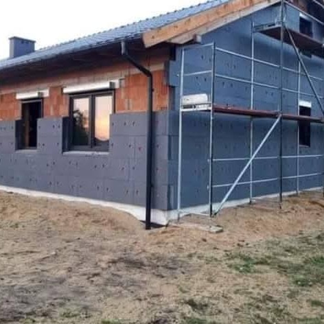 Ocieplanie budynku styropianem grafitowym, widoczne rusztowanie i fragment ściany z cegły. Prace termoizolacyjne w toku, etap przed nałożeniem tynku.