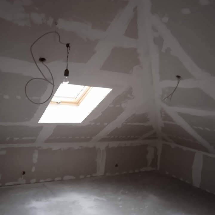 Poddasze po szpachlowaniu łączeń płyt gipsowych, widoczne okno dachowe i wystające przewody oświetleniowe. Ściany i sufit przygotowane do malowania.