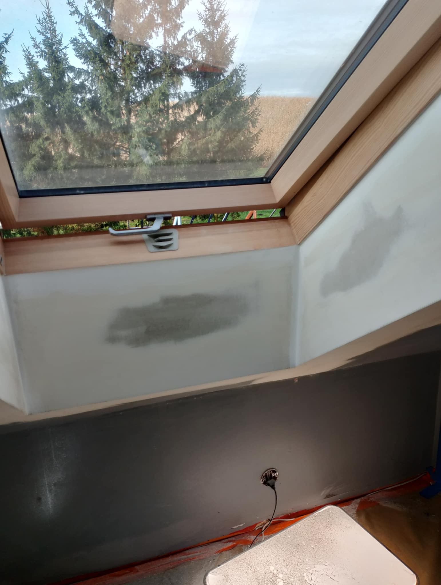 Ściana poddasza ze skośnym oknem dachowym w trakcie szpachlowania. Widoczne niedoskonałości i plamy po szpachli. Na dole ściana pomalowana na szaro.