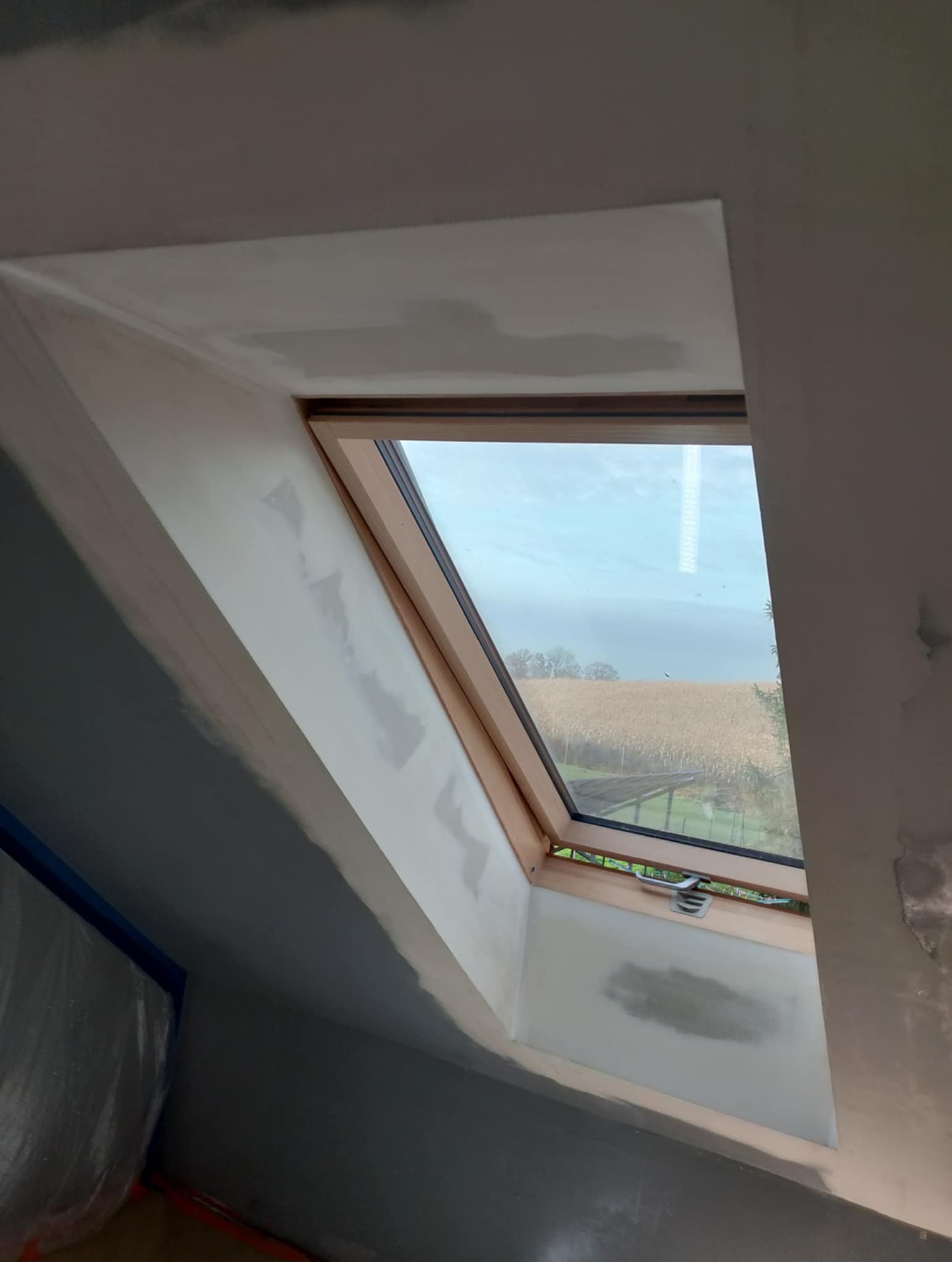 Poddasze z oknem dachowym po szpachlowaniu, widoczne ślady po łatach gipsowych. Okno drewniane, na zewnątrz widok na pole. Ściany w trakcie malowania.