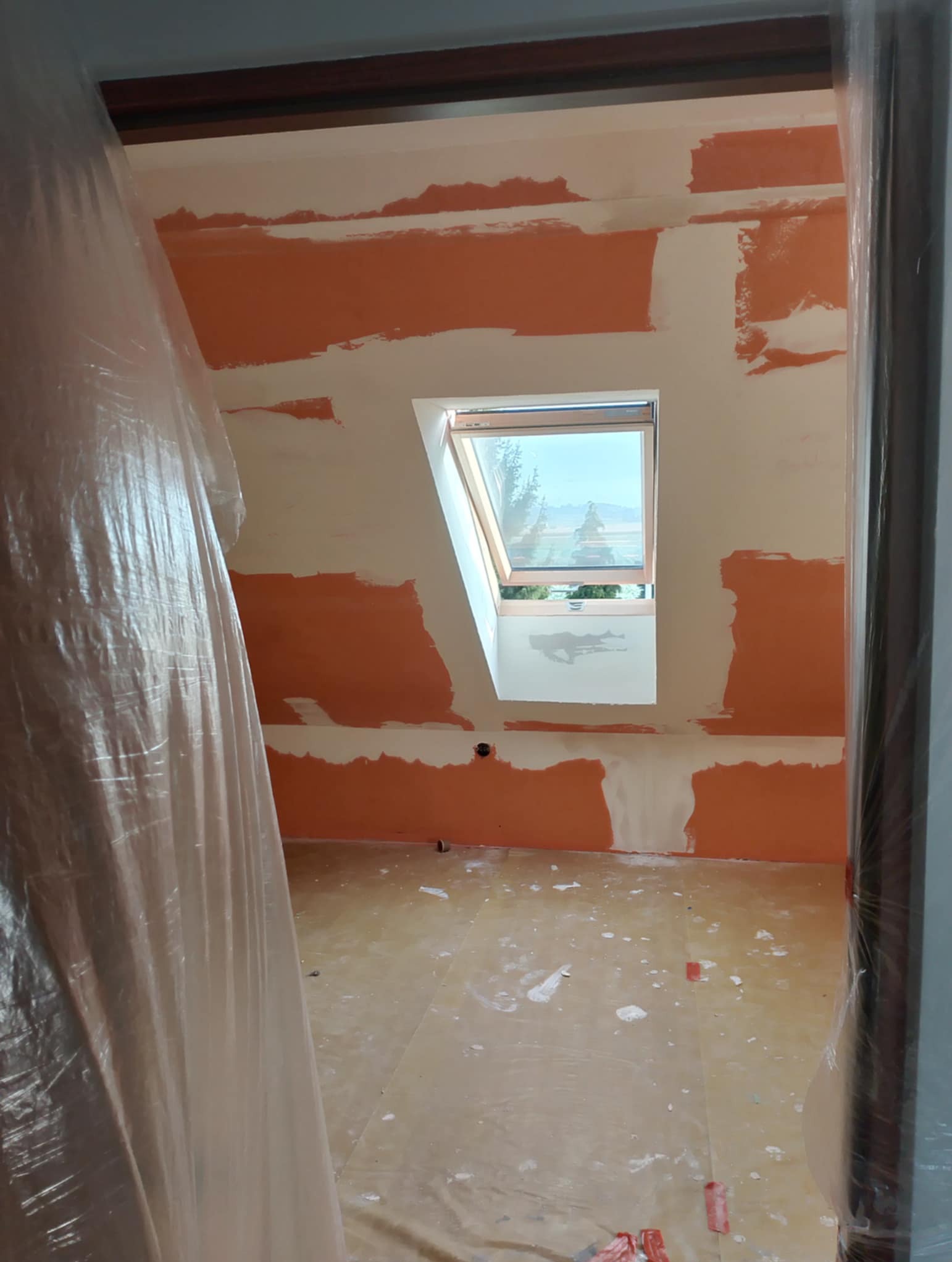 Pokój na poddaszu w trakcie szpachlowania, zabezpieczony folią malarską. Widoczne fragmenty pomarańczowej farby i okno dachowe.