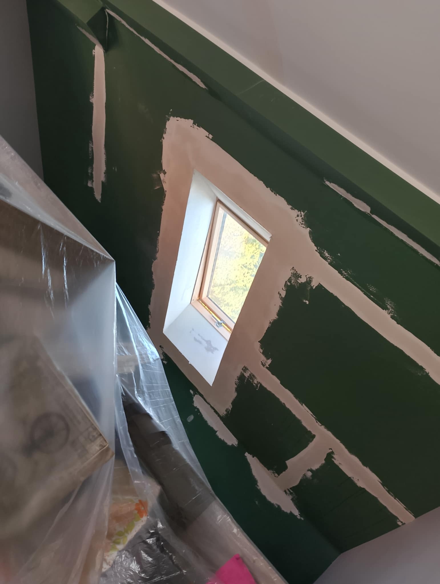 Skośna ściana w kolorze butelkowej zieleni z oknem dachowym w trakcie szpachlowania. Widoczne paski szpachli wokół okna i na łączeniach płyt. Część mebli zabezpieczona folią.
