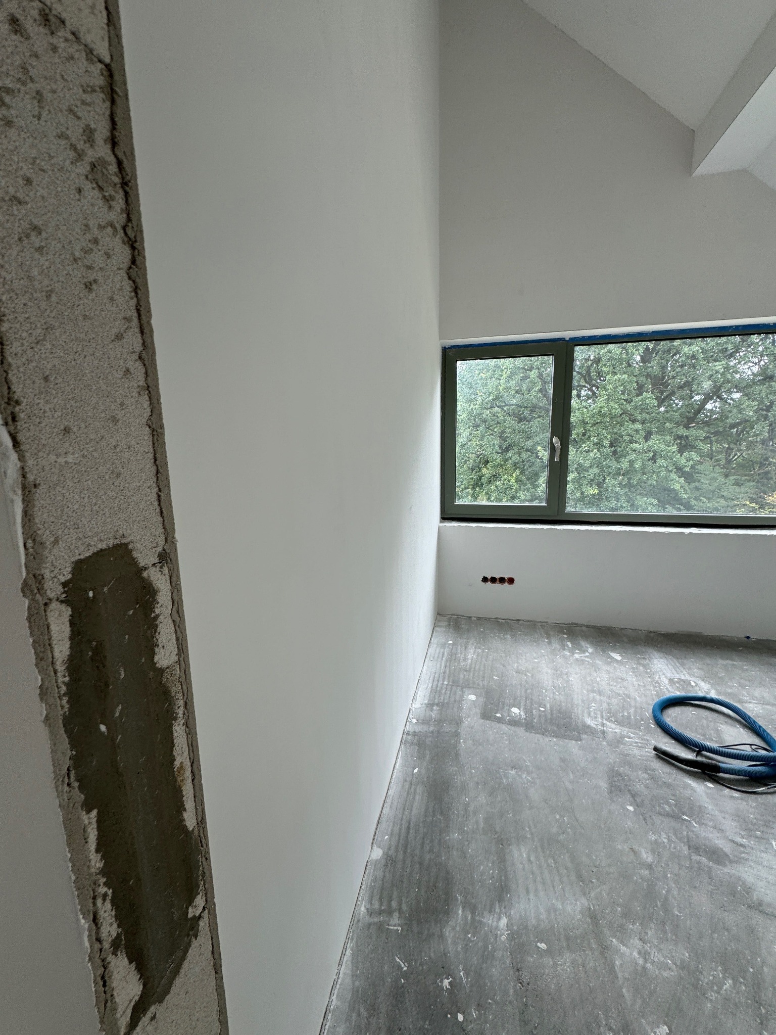 Wnętrze w trakcie prac wykończeniowych: gładkie, białe ściany, surowa podłoga, okno z widokiem na zieleń. Widoczny fragment ściany w stanie surowym i wąż od odkurzacza.