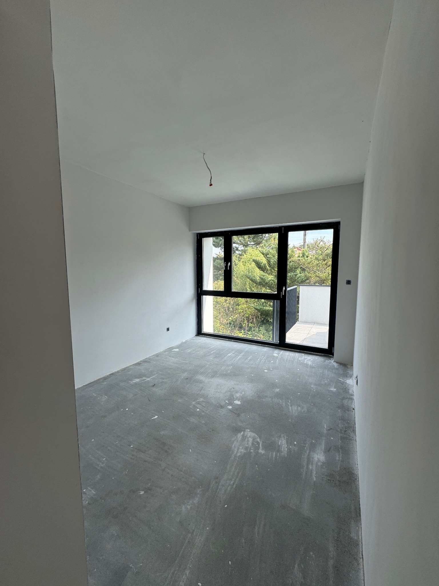 Surowe wnętrze z dużym oknem balkonowym, szare ściany i podłoga w trakcie przygotowania do wykończenia. Widok na zieleń z balkonu.