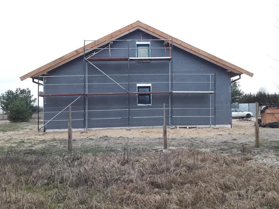 Ocieplony budynek z szarego bloczka, z drewnianą konstrukcją dachu i rusztowaniem. Widoczne okna i otoczenie budynku: trawa, drzewa, samochód.