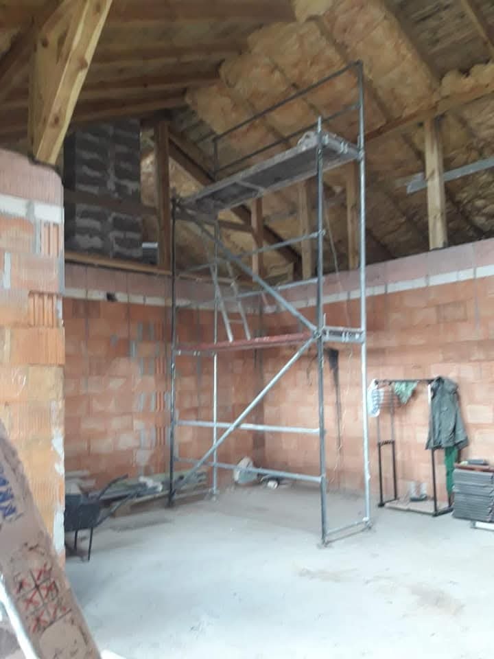 Wnętrze poddasza w trakcie prac wykończeniowych. Widoczne rusztowanie, ściany z cegły, izolacja dachu i elementy konstrukcji drewnianej. Etap przygotowania do dalszych prac.