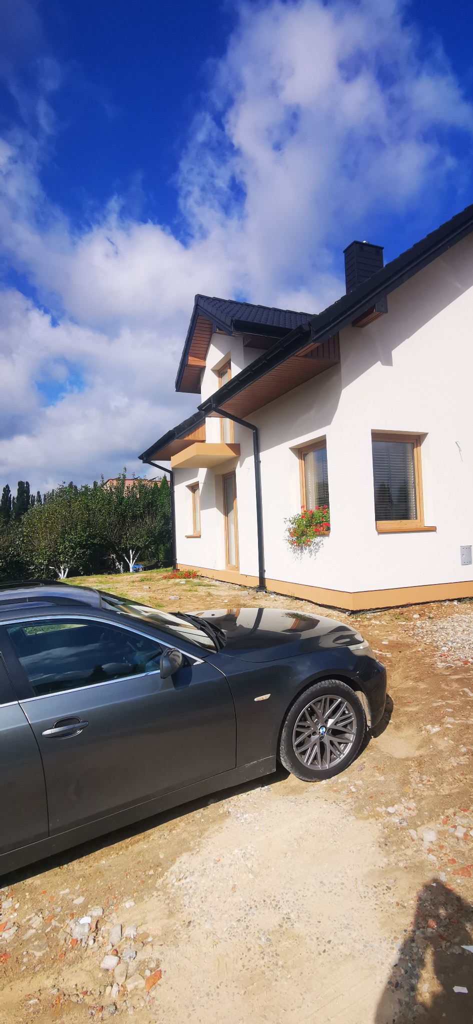 Biała elewacja domu jednorodzinnego z brązowymi oknami i czarnym dachem, na tle błękitnego nieba z chmurami. Na pierwszym planie zaparkowany szary samochód.