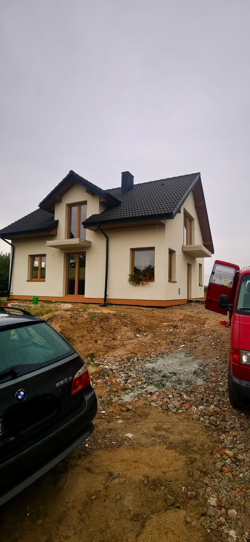 Nowo wybudowany dom jednorodzinny z ciemną dachówką i jasną elewacją, z widocznymi oknami z drewnianymi ramami i balkonem. Na pierwszym planie samochody.