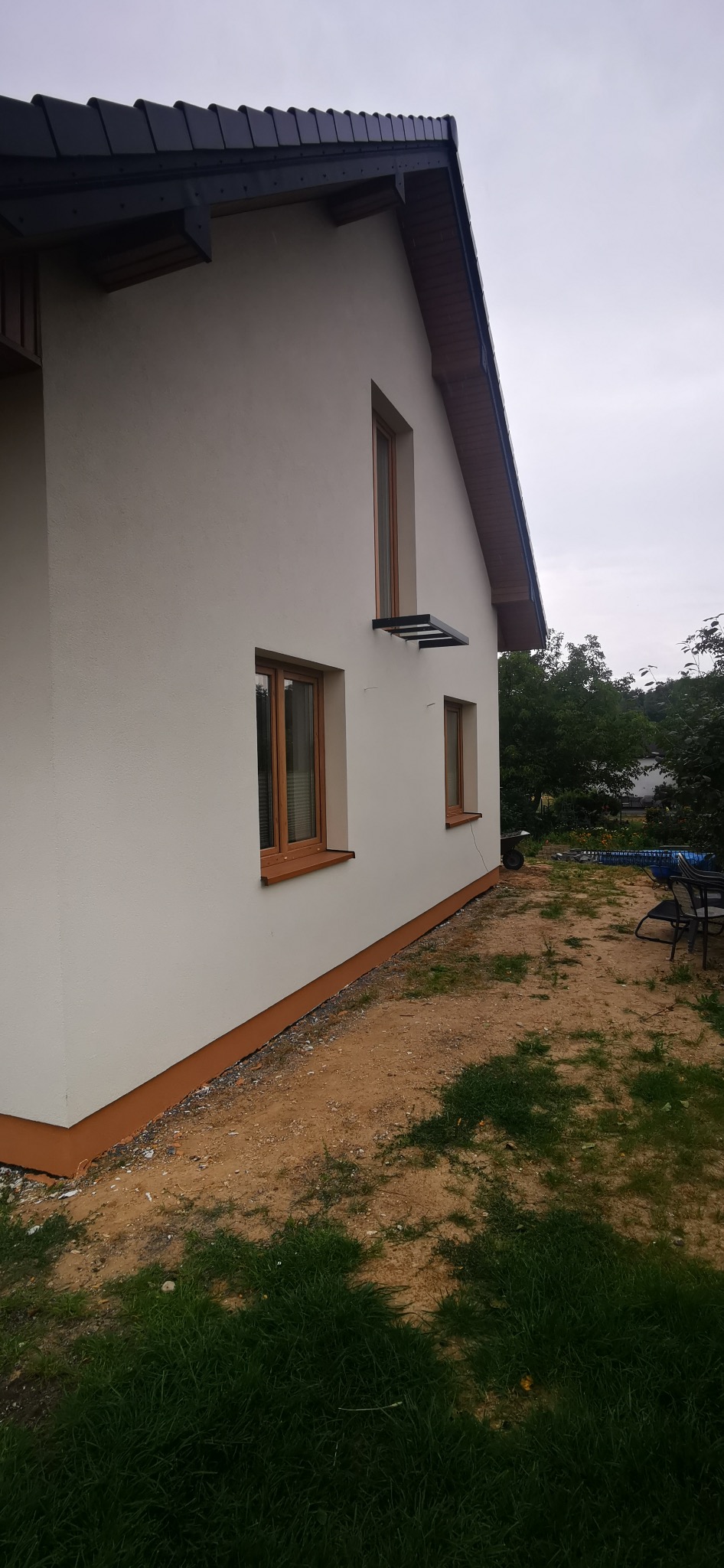 Elewacja domu jednorodzinnego z brązowymi oknami i podbitką, widok z boku na tle zieleni i pochmurnego nieba. Widoczny fragment dachu z ciemną dachówką.