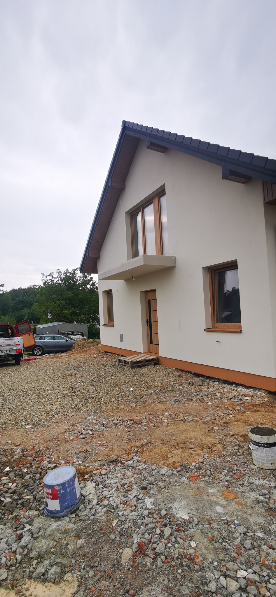 Elewacja domu jednorodzinnego z drewnianą stolarką okienną i drzwiami, widok z zewnątrz na tle pochmurnego nieba i zieleni. Teren wokół domu w trakcie prac wykończeniowych.