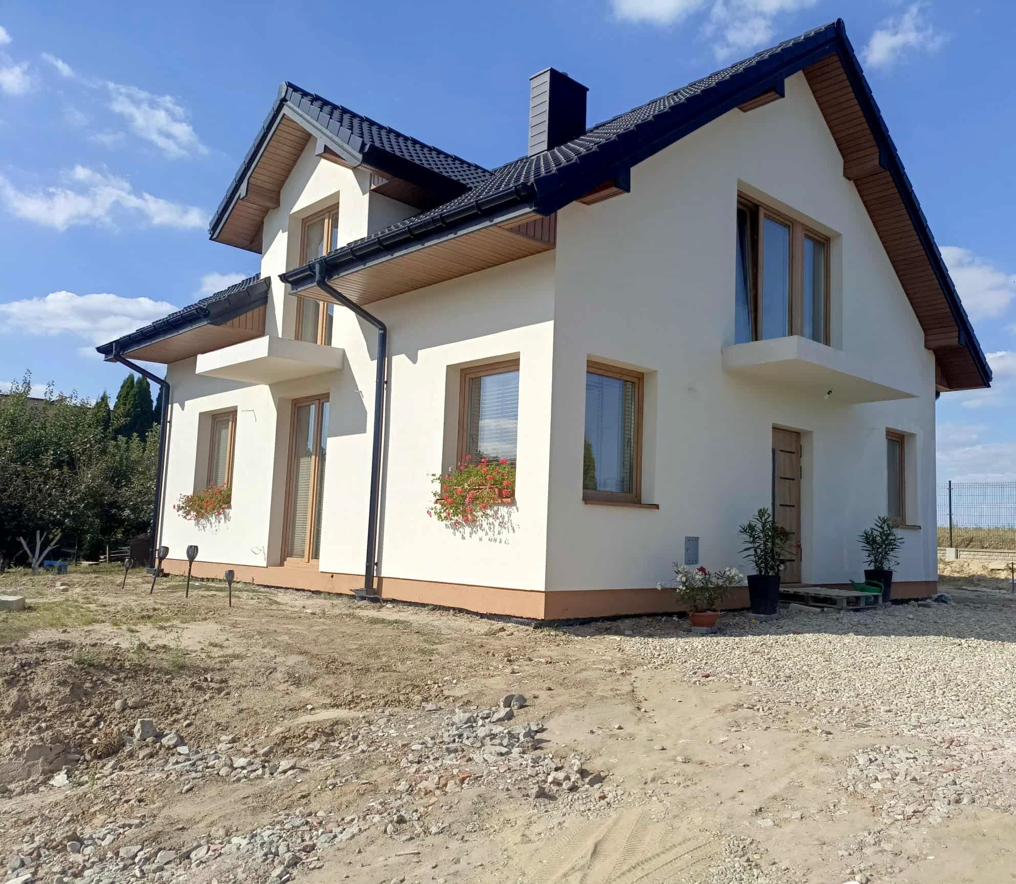 Nowoczesny, dwukondygnacyjny dom z białymi ścianami, ciemnym dachem i drewnianymi oknami. Widoczne kwiaty w skrzynkach okiennych i rośliny w donicach przy wejściu.