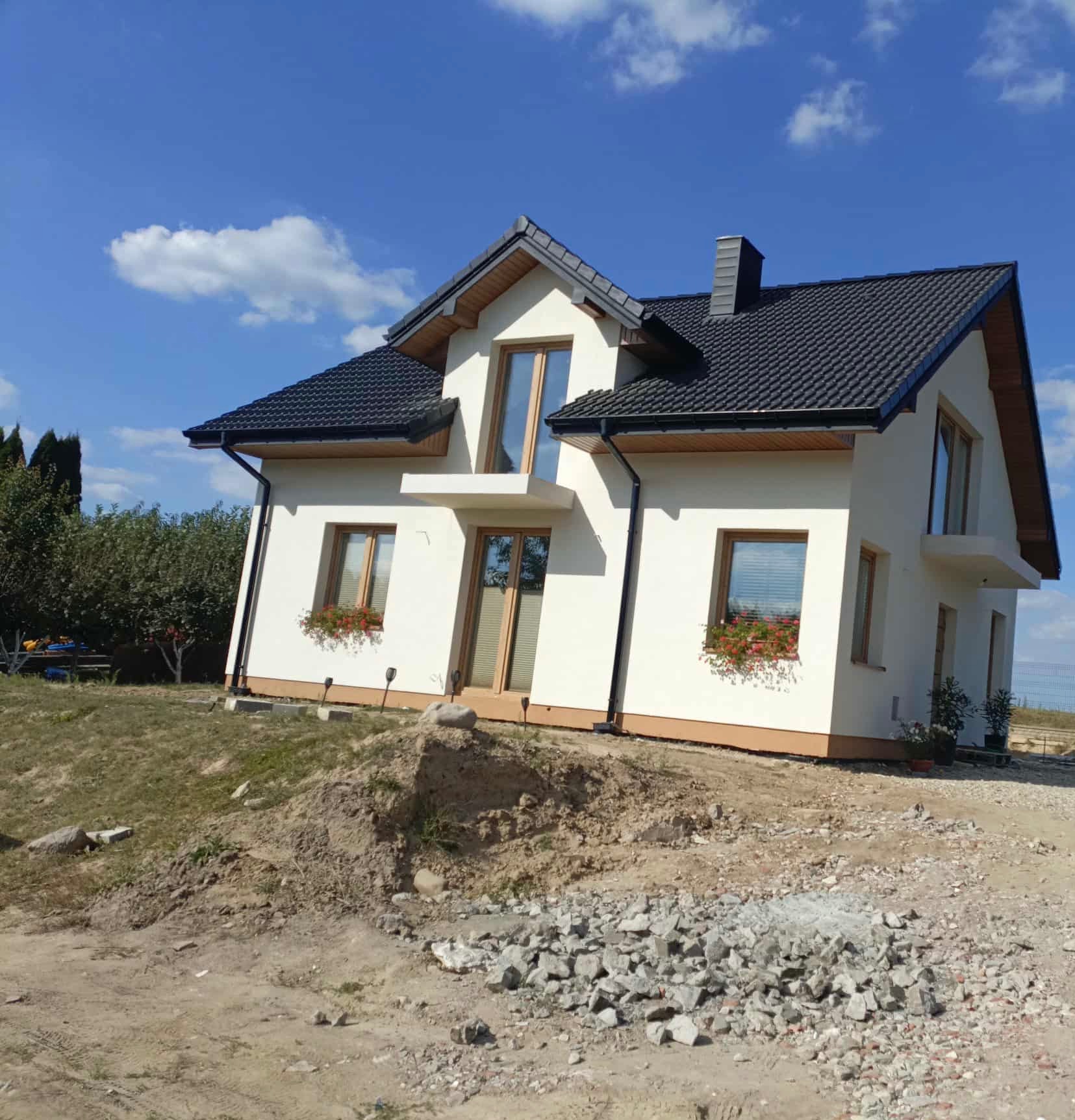 Biały dom z ciemnym dachem na wzgórzu, widok z dołu, z oknami z kwiatami i elementami wykończeniowymi w kolorze drewna, na pierwszym planie gruz i ziemia.