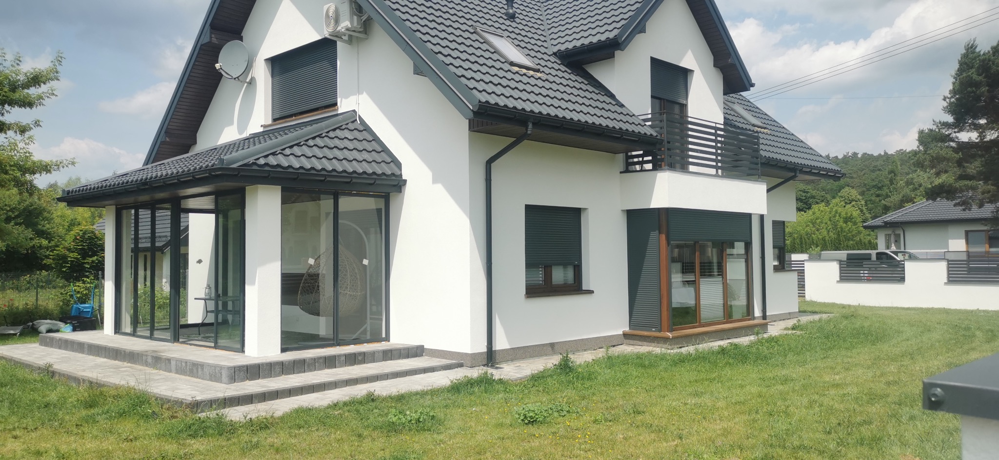 Biały dom jednorodzinny z ciemną dachówką i roletami zewnętrznymi, widok z zewnątrz na przeszkloną werandę i balkon z metalową balustradą, otoczony zielonym trawnikiem.