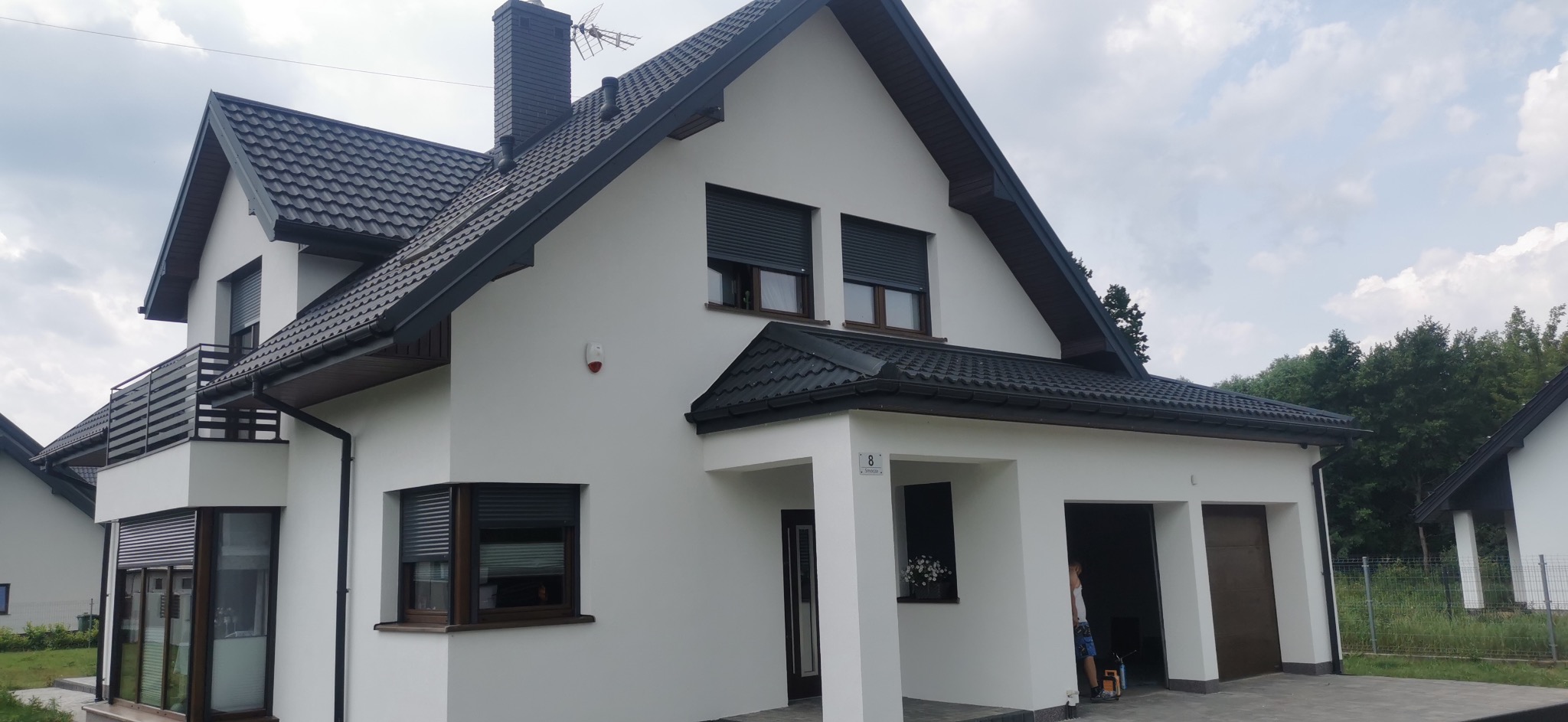 Biały dom jednorodzinny z ciemną dachówką i roletami zewnętrznymi, widok z zewnątrz, świeżo po malowaniu elewacji, z numerem domu przy wejściu.