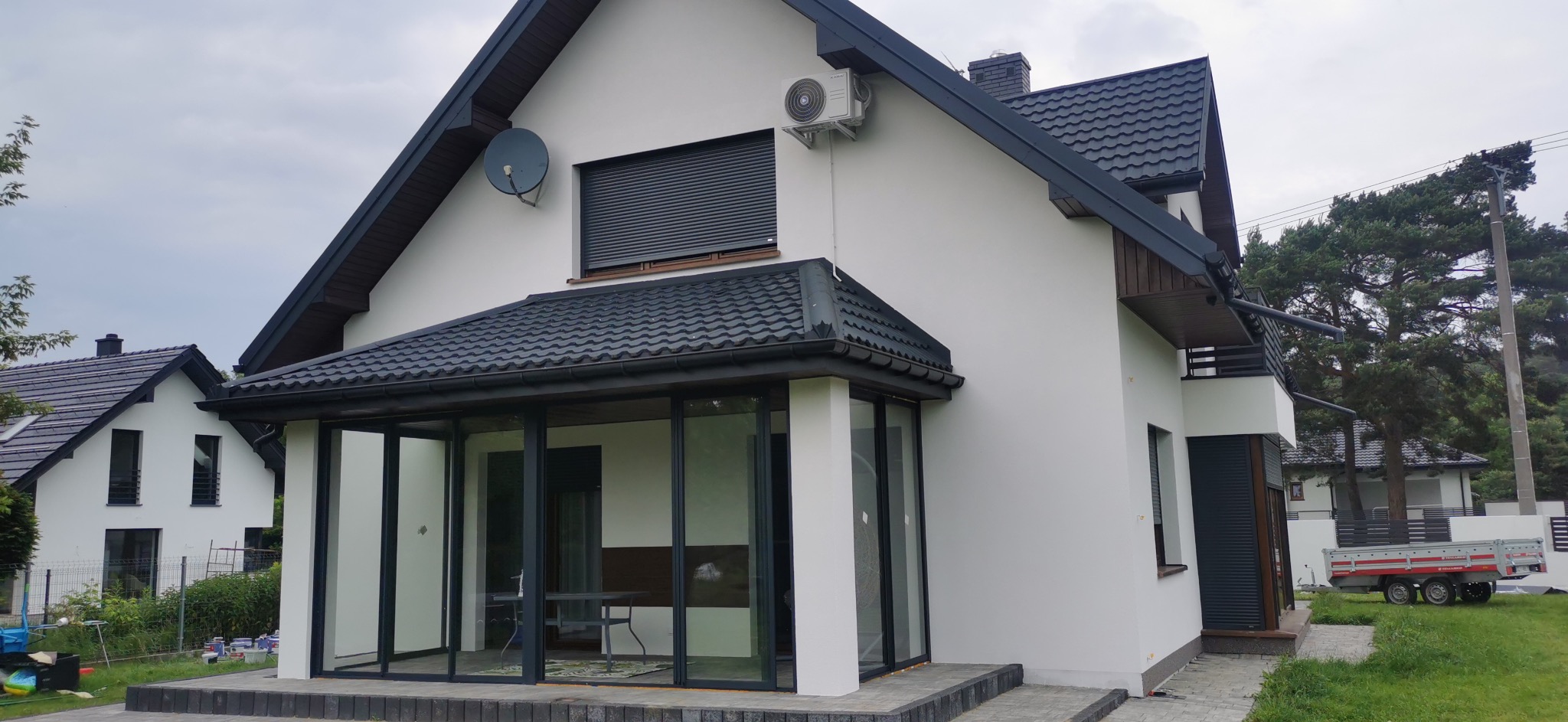Elewacja domu jednorodzinnego z przeszklonym tarasem i roletami zewnętrznymi. Widoczny klimatyzator, antena satelitarna i fragment ogrodu z przyczepą.