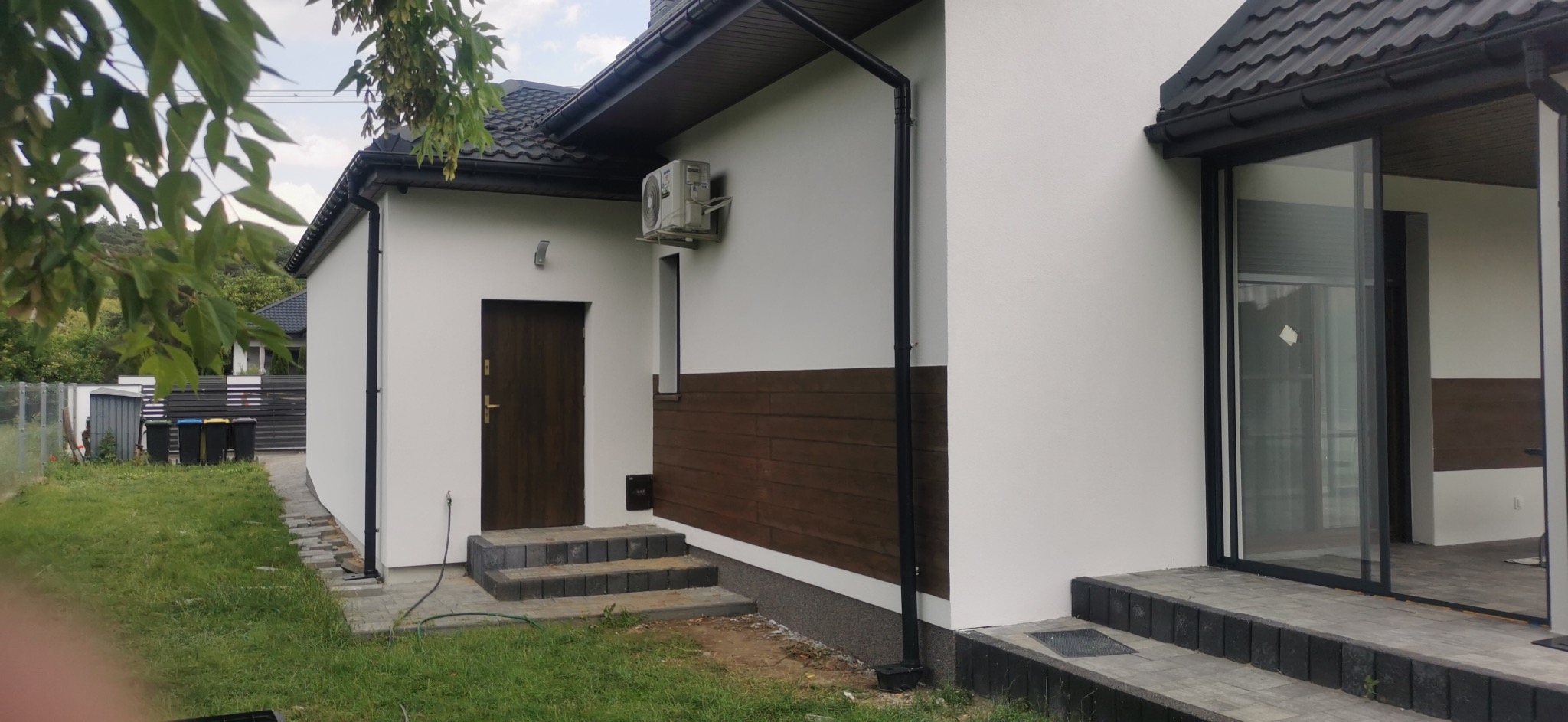 Biała elewacja domu z brązowymi akcentami: drewniana boazeria, drzwi i okna. Widoczny klimatyzator, rynna i schody wejściowe. Tło stanowi zieleń drzew.