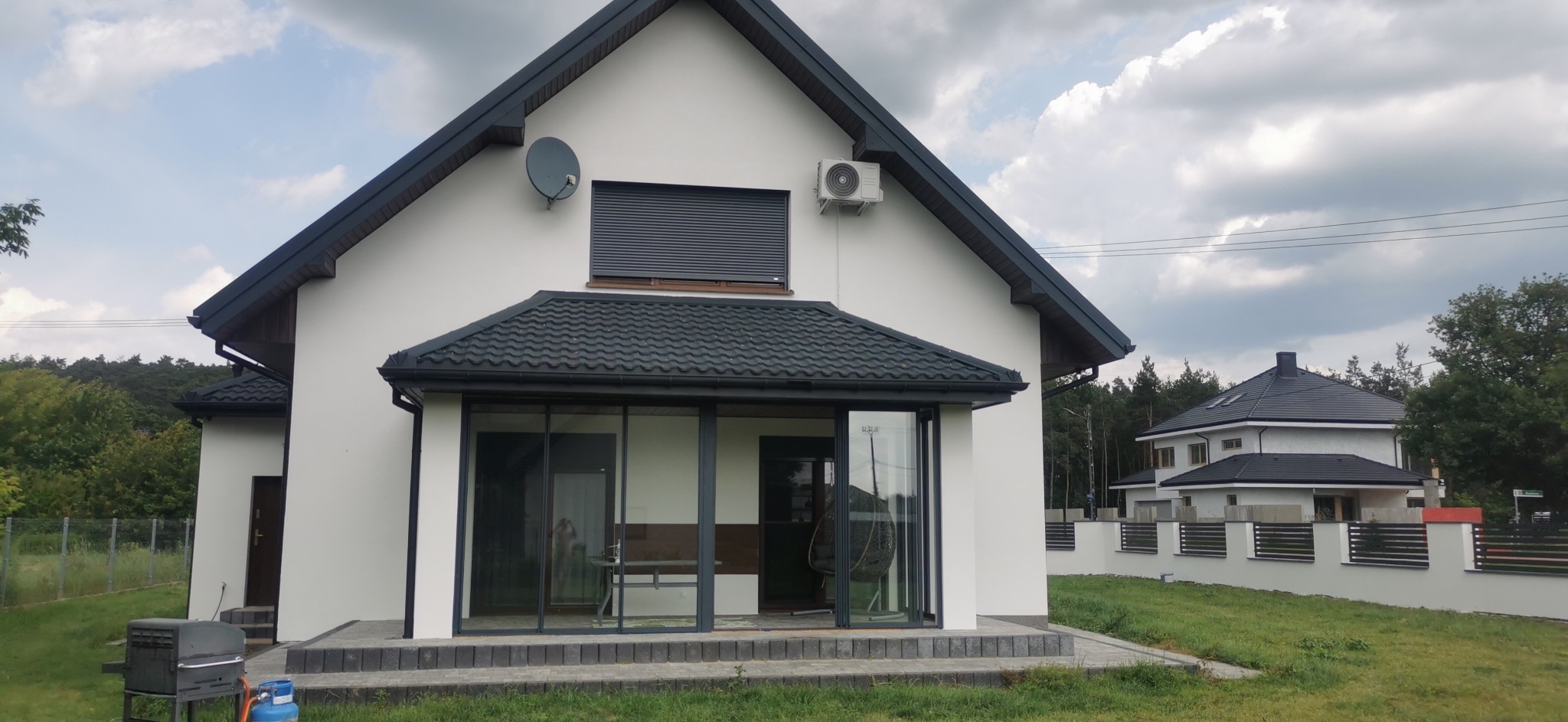 Biały dom z ciemną dachówką i dużymi oknami, widok z zewnątrz w słoneczny dzień. Widoczna antena satelitarna i klimatyzator na elewacji. Nowoczesny styl.