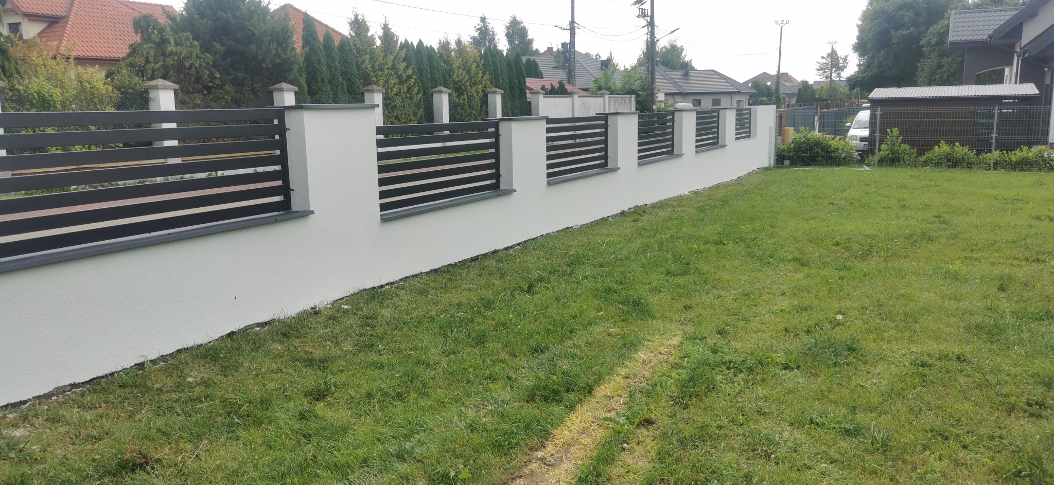 Nowoczesne ogrodzenie posesji z białymi słupkami i poziomymi, grafitowymi przęsłami, widok z zewnątrz na tle zielonego trawnika i zabudowań.