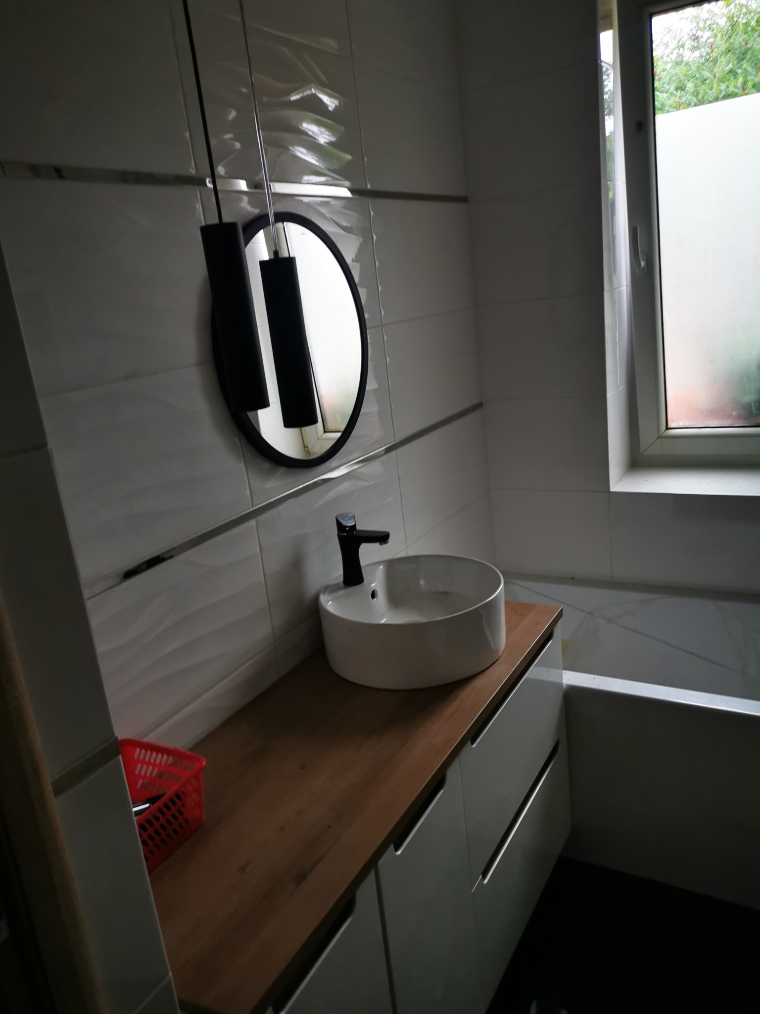 Nowoczesna łazienka z umywalką nablatową, drewnianym blatem i okrągłym lustrem. Białe płytki z subtelnym wzorem i minimalistyczna lampa wisząca.