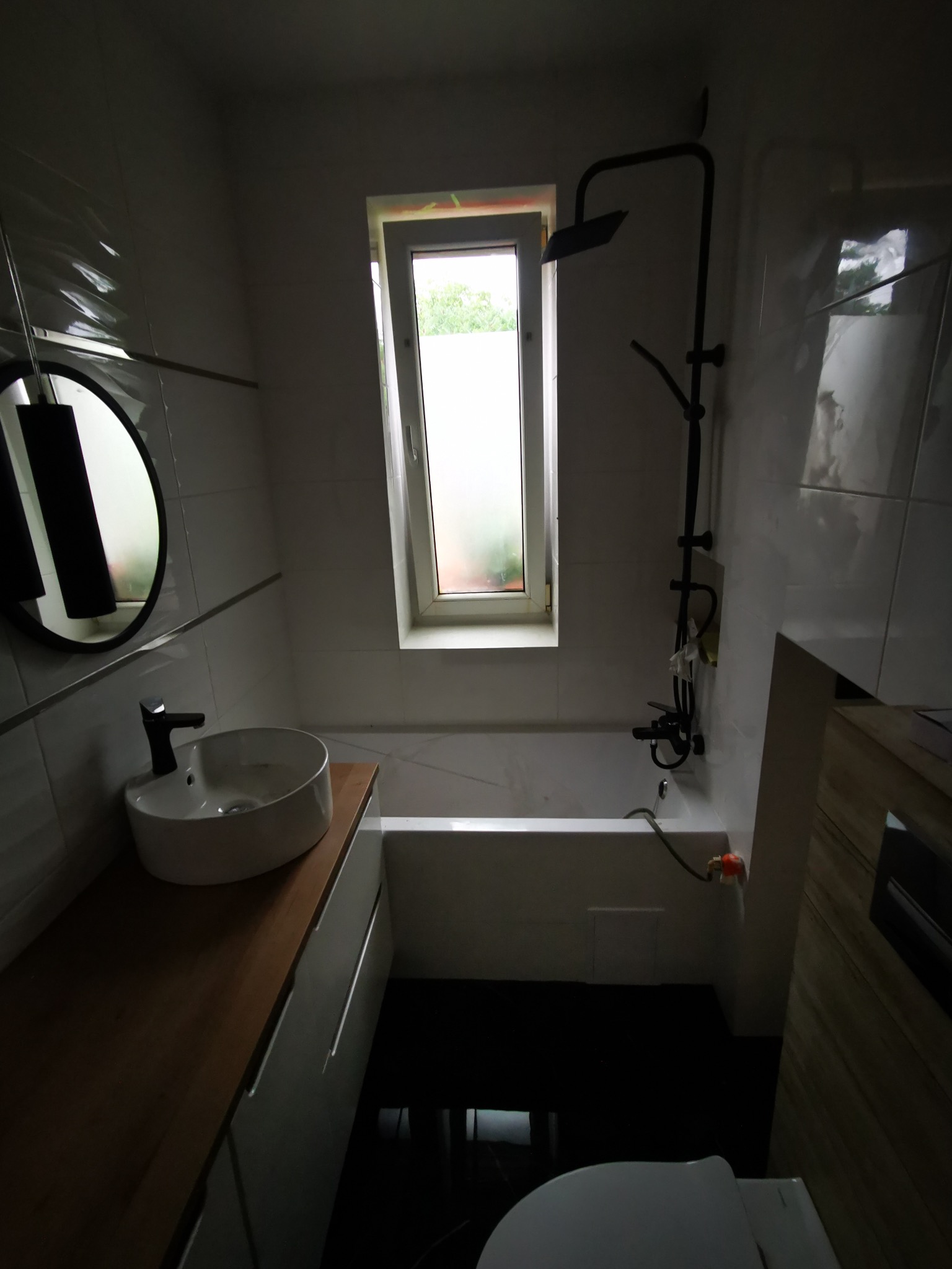 Nowoczesna łazienka z owalną umywalką na drewnianym blacie, czarną armaturą i wąskim oknem. Białe płytki z subtelnym wzorem na ścianach.