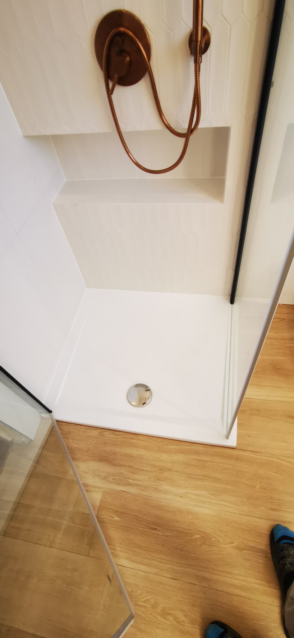 Nowoczesna kabina prysznicowa z białą brodzikiem i armaturą w kolorze miedzi. Ściany wyłożone białymi, podłużnymi płytkami. Widoczny fragment drewnianej podłogi.