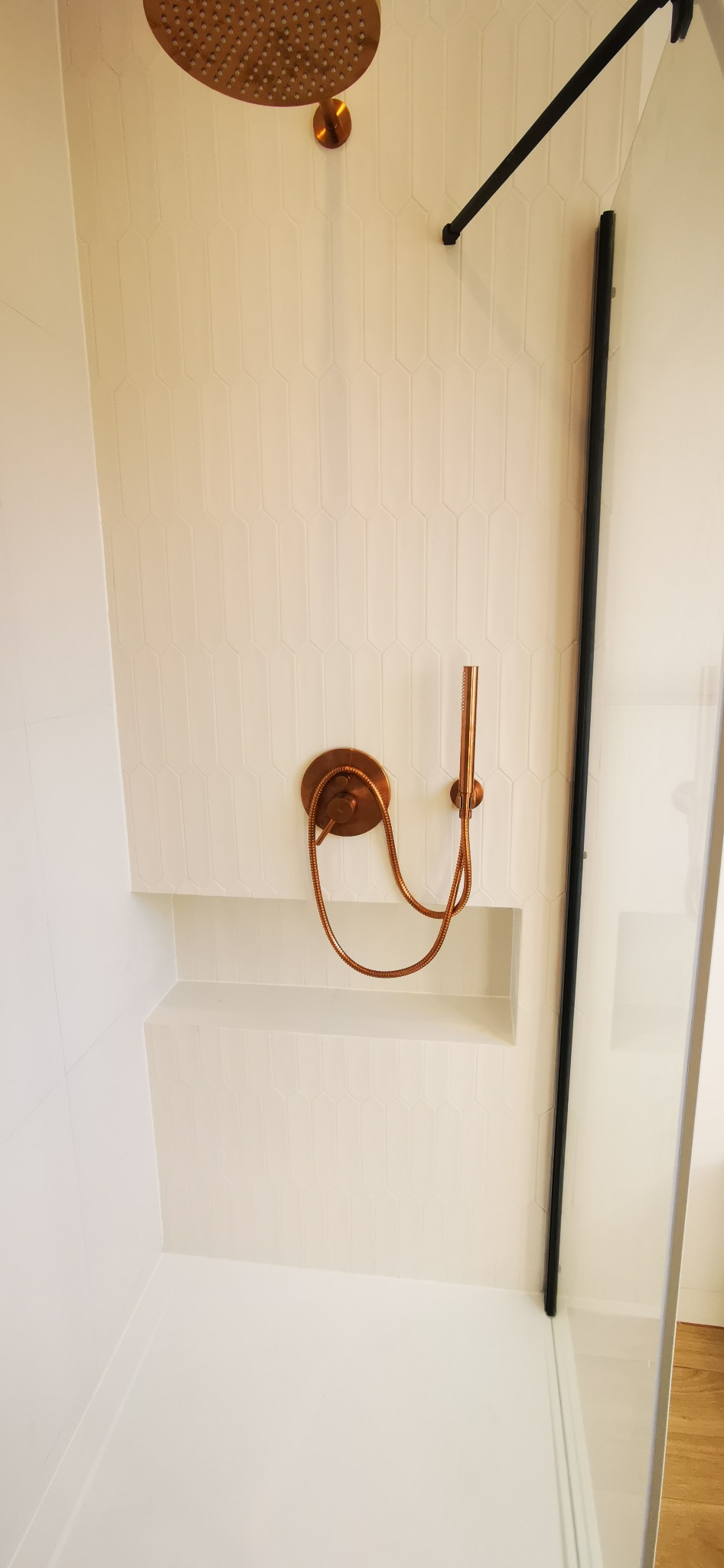 Nowoczesna kabina prysznicowa z armaturą w kolorze miedzi i białymi, podłużnymi płytkami. Widoczna deszczownica, bateria i wnęka na kosmetyki.