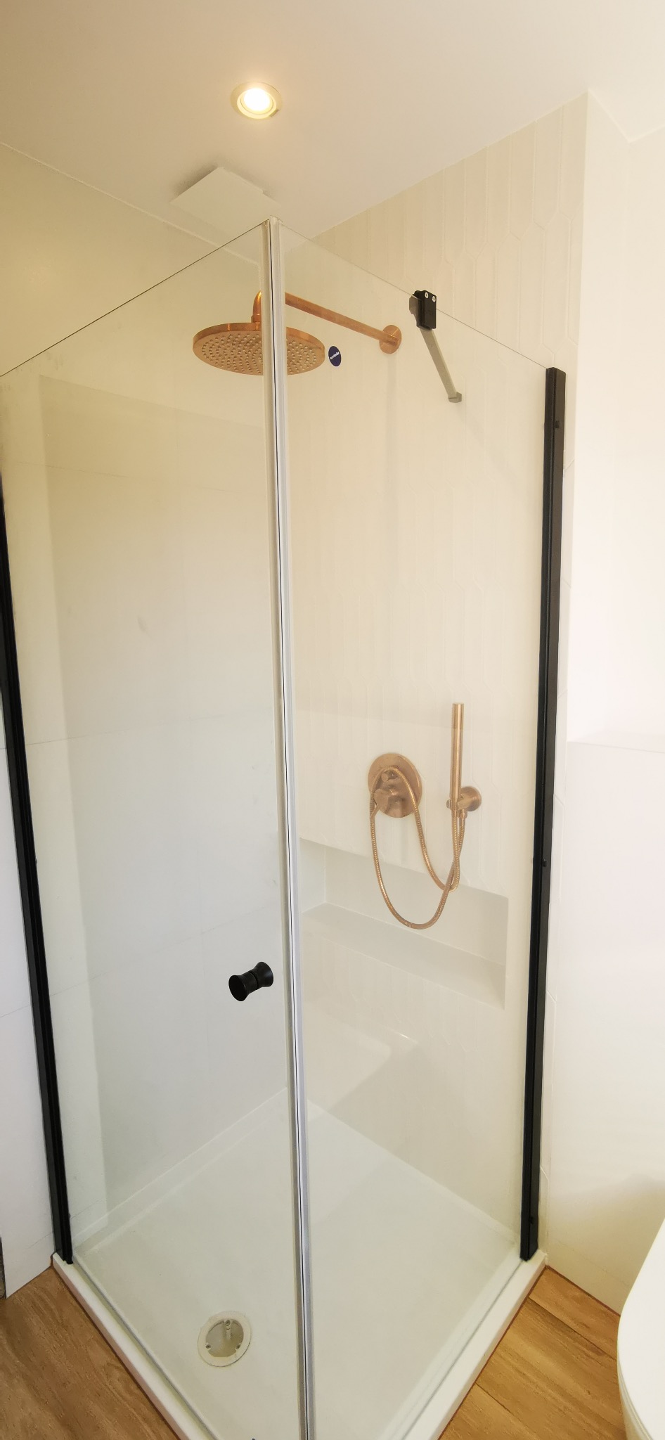 Nowoczesna kabina prysznicowa z deszczownicą w odcieniach miedzi, czarne ramy, białe płytki heksagonalne i drewnopodobna podłoga. Minimalistyczny design łazienki.