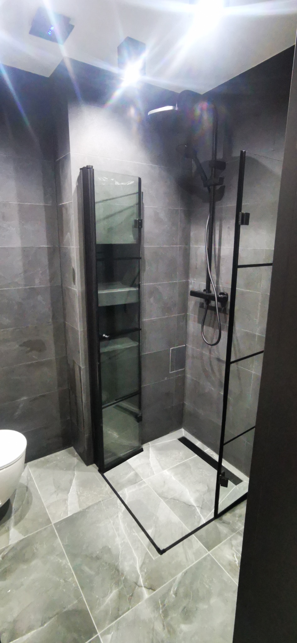 Nowoczesna łazienka z szarymi płytkami, kabiną prysznicową typu walk-in z czarnymi ramami i szklaną półką. Minimalistyczny design, oświetlenie sufitowe.