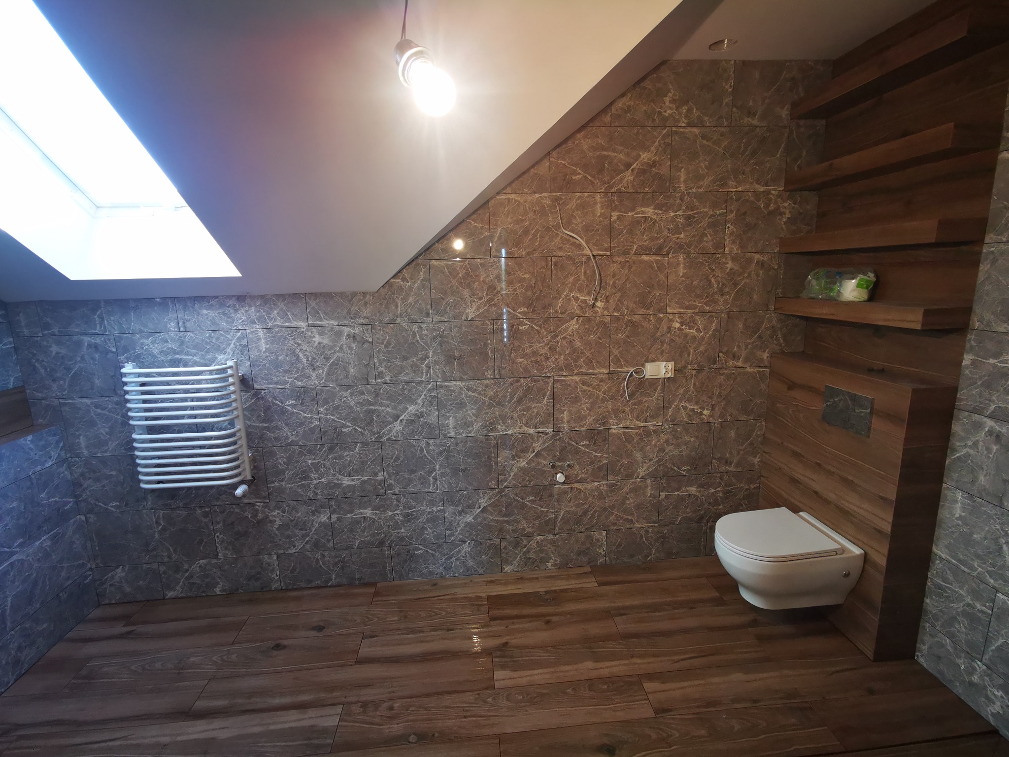 Ukończona łazienka na poddaszu z marmurowymi płytkami, drewnianą podłogą i grzejnikiem drabinkowym. Widoczne skosy dachu i okno dachowe.