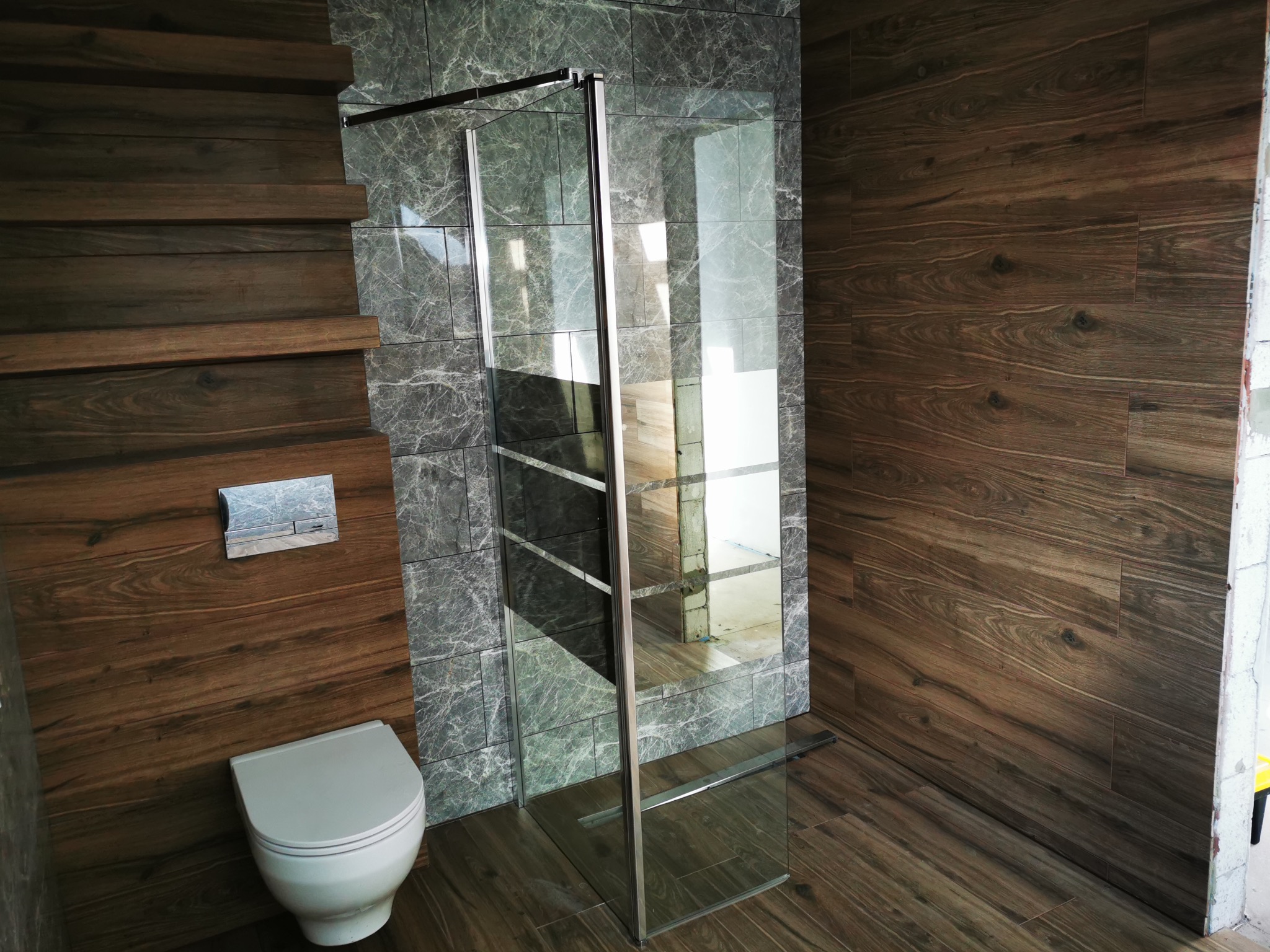 Nowoczesna łazienka z kabiną prysznicową typu walk-in, wykończona płytkami imitującymi drewno i marmur. Widoczny podtynkowy system WC i minimalistyczna armatura.