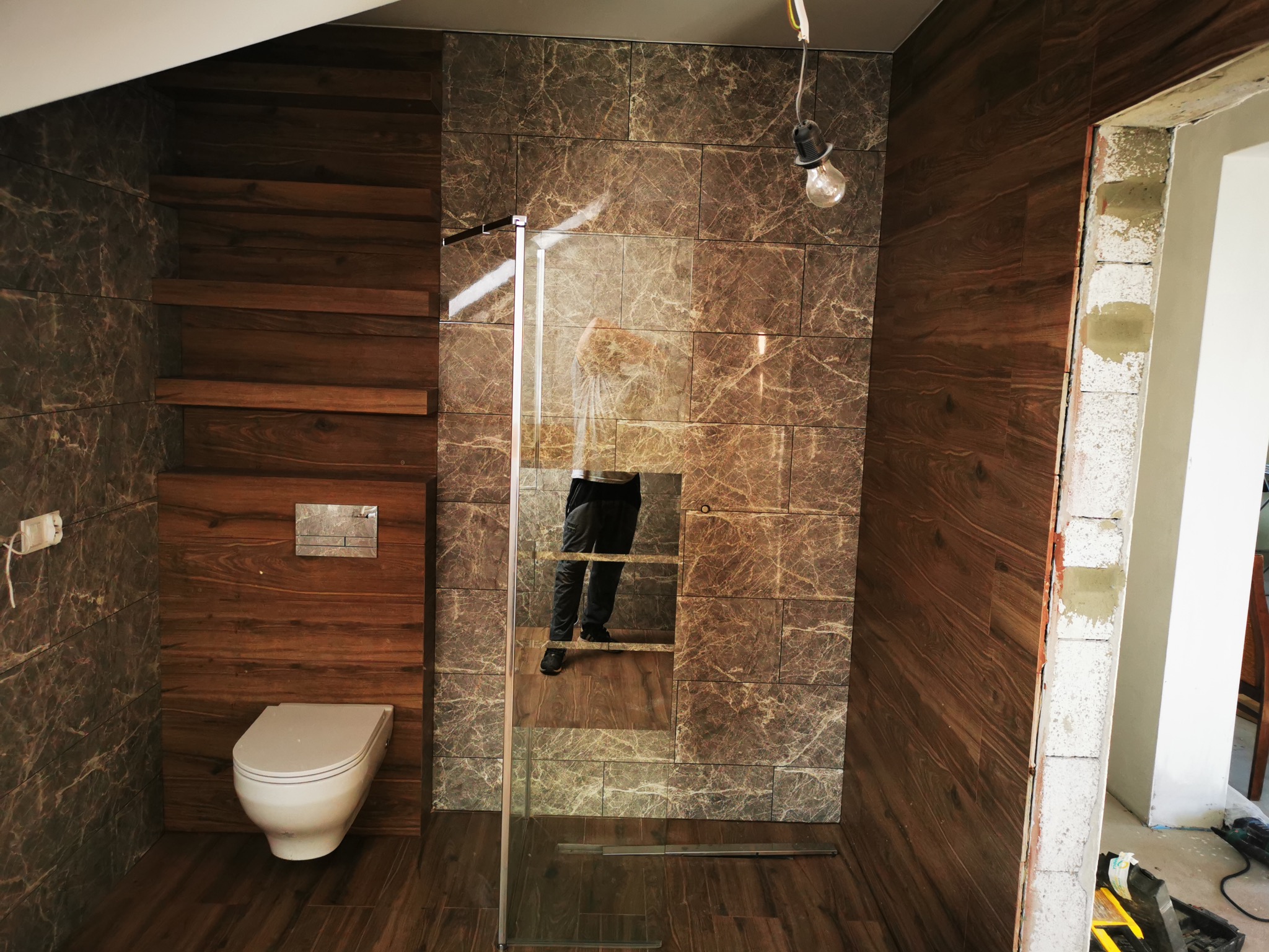 Wykończona łazienka z brązowymi płytkami imitującymi drewno, kabiną prysznicową ze szkła i wiszącą toaletą. Widoczne niedokończone obramowanie drzwi.