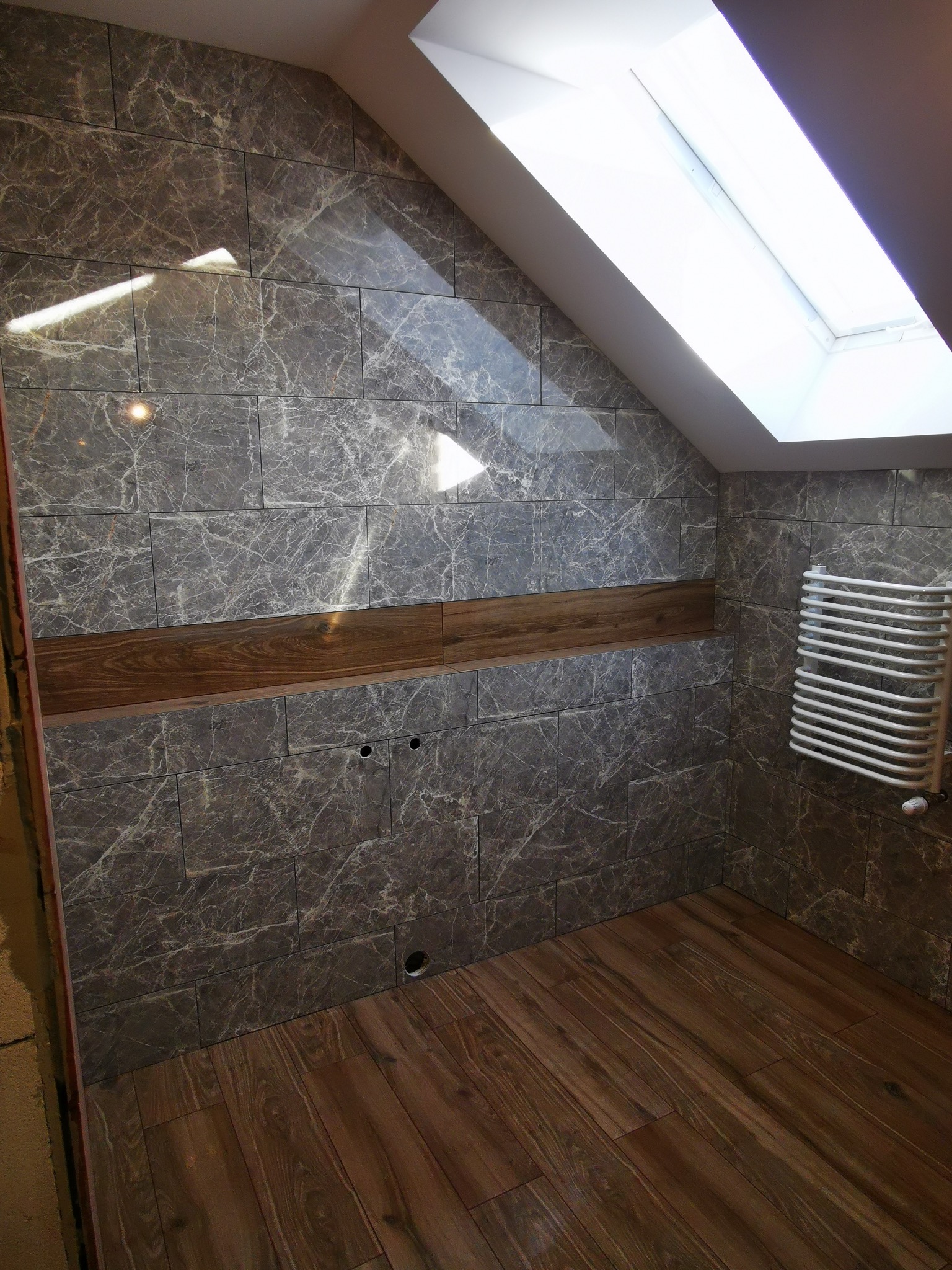 Wykończona łazienka na poddaszu z szarymi płytkami imitującymi marmur, drewnianą listwą dekoracyjną i grzejnikiem drabinkowym. Widoczne okno dachowe i drewniana podłoga.